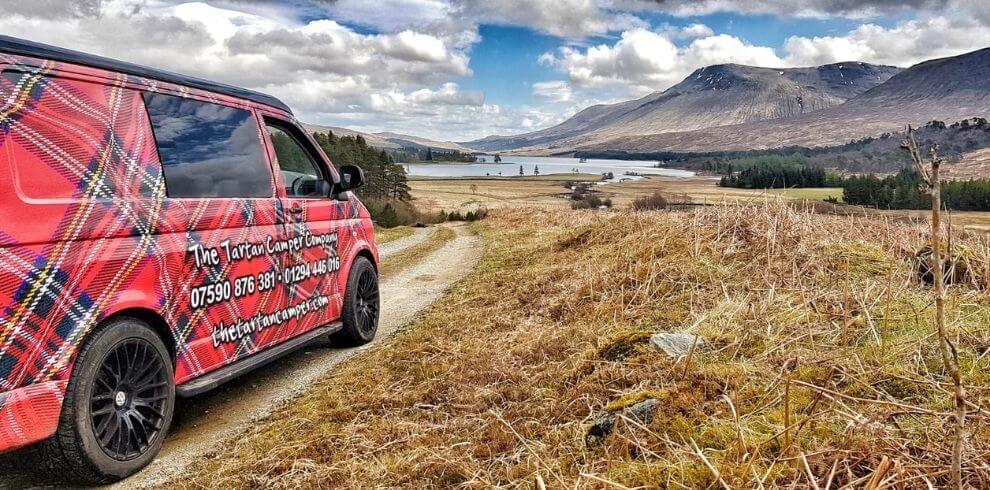 a tartan campervan driving towards a loch and glen