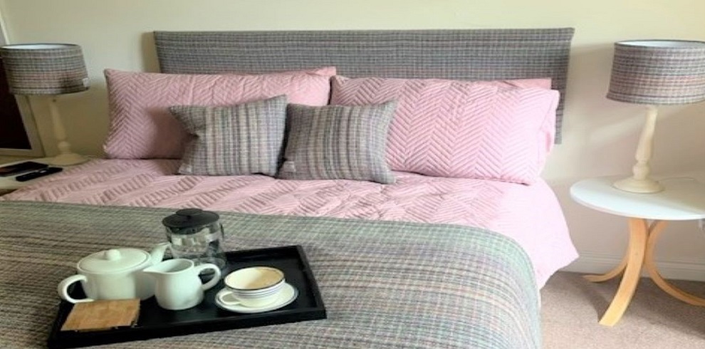 A double bed with pink and grey covers with a tray sitting on the bed with cups of coffee.