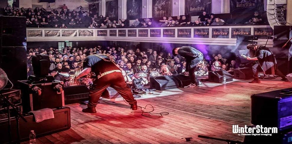 An performer playing bass/guitar on stage with the audence looking on in the background.