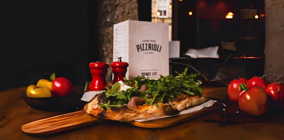 An image of a homemade pizza placed on a table. There is a menu in the background that says 'Pizzaioli' on it.