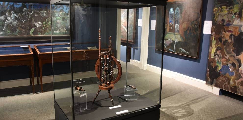 Spinning wheel in glass case surrounded by large artwork.
