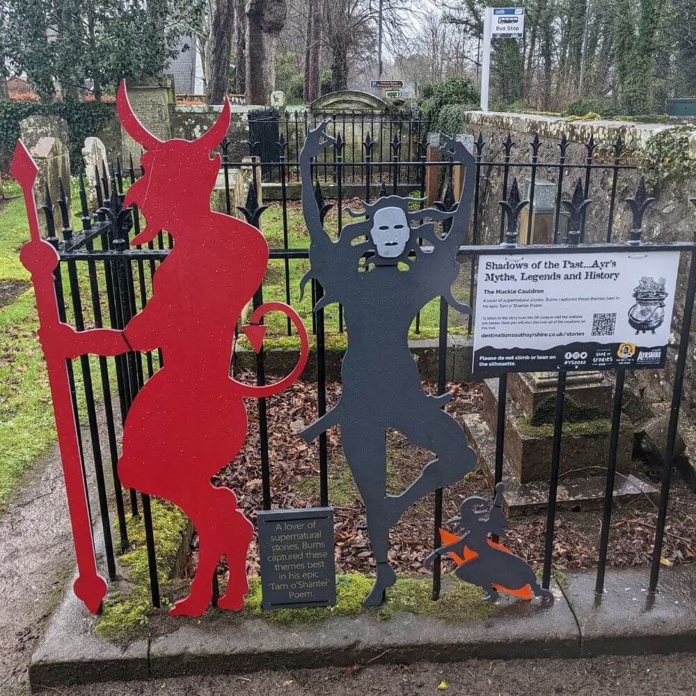 A wooden silhouette of a red devil and dancing ghoul against a metal fence.