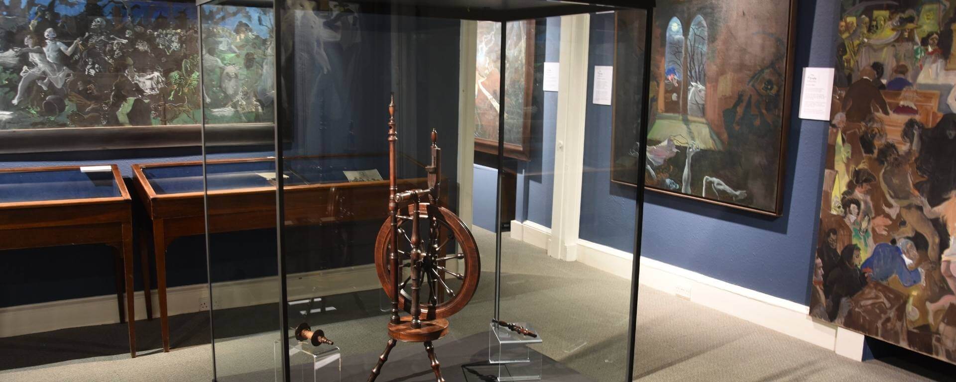 Spinning wheel in glass case surrounded by large artwork.