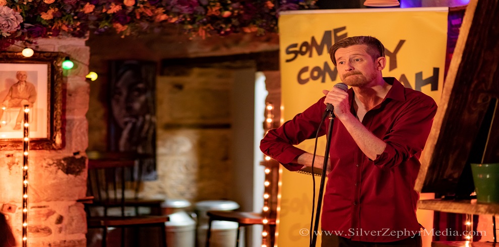 A picture of a male standing on stage holding a microphone. There is a yellow sign behinf him that says 'Some Comedy Yeah'.