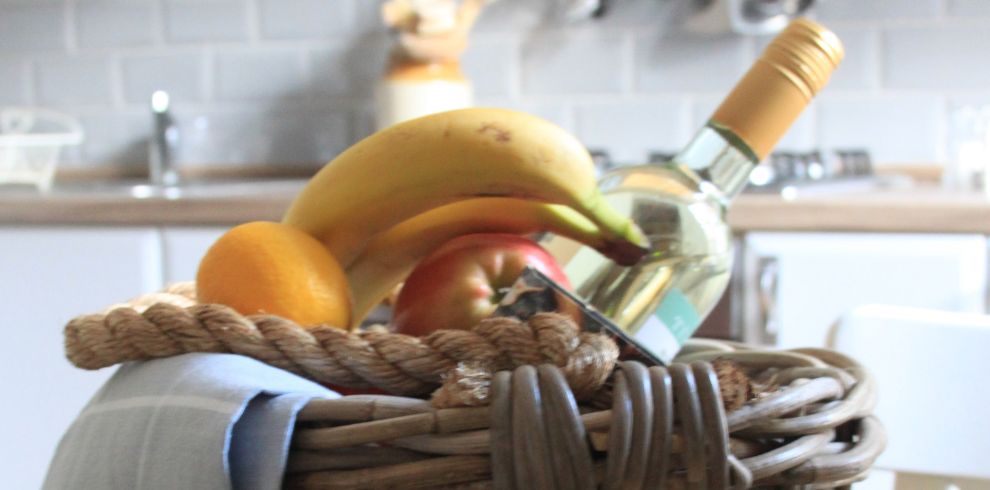Close up of a whicker basket containing fruit and a bottle of wine in a kitchen.