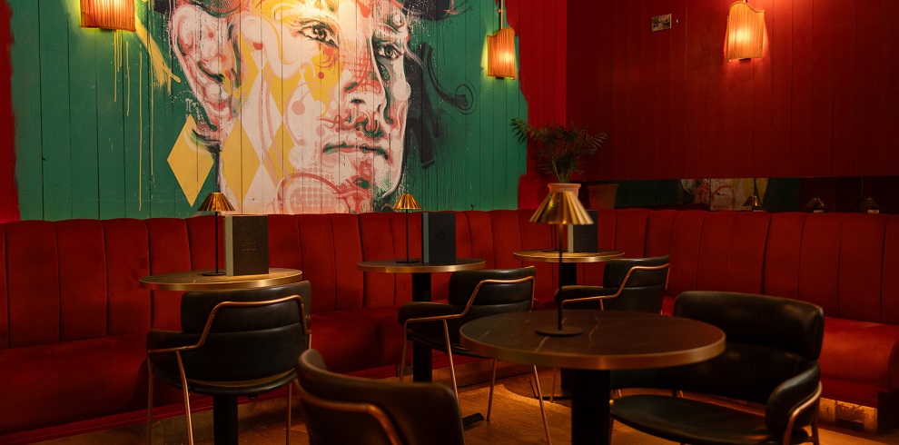 This image showcases a stylish and cosy lounge area with modern decor. The space features a vibrant mural of a person's face with colorful geometric patterns on a teal and red background. The furniture includes black leather chairs and a red upholstered banquette lining the walls. The room is warmly lit by pendant lights with golden shades and spherical white lamps hanging from the ceiling. A projector screen is visible on one side, suggesting the room is multifunctional for events or entertainment. The wooden floor adds a natural touch to the overall contemporary design.