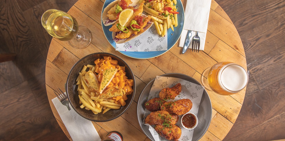 This image captures a top-down view of a wooden table set with appetising food and drinks. The table features three dishes: a plate of fries topped with melted cheese and garnished with herbs, a plate of crispy fried chicken wings served with a dipping sauce, and a sandwich with fries, garnished with lemon and peppers. Two beverages accompany the meal — a glass of white wine and a pint of beer. A hand reaches in to pick up the sandwich, adding a dynamic touch to the composition. The setup evokes a casual, inviting dining atmosphere.