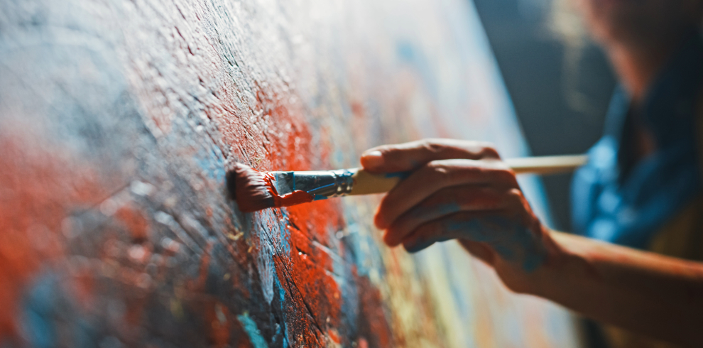 Image of a hand holding a paintbrush, painting on a canvas.