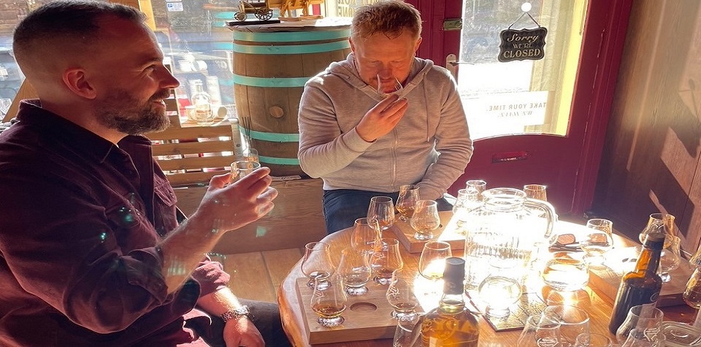 Two people sitting at a table enjoying whisky. One person is holding the glass up to their nose to have a smell.