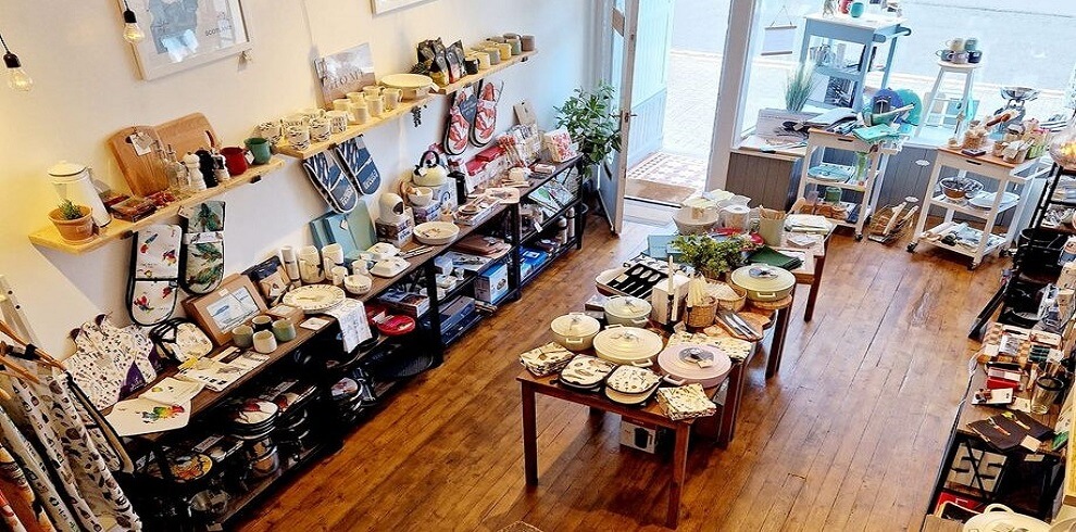 The photo shows the interior of Bay & Berry kitchenware shop. Tables are neatly arranged with various kitchen items on display. The tables are covered with colorful plates, bowls, utensils, and cookware. The shop has a cosy and inviting atmosphere, with wooden shelves lining the walls, also filled with kitchen products. Natural light streams in from large windows, enhancing the vibrant colors of the merchandise. The overall layout is organized, making it easy for customers to browse the diverse selection of kitchenware.