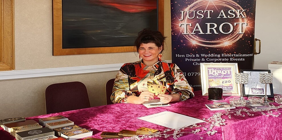 A smiling lady sits at a table with a set of tarot cards spread out. Beside her, a sign reads "Just Ask Tarot.