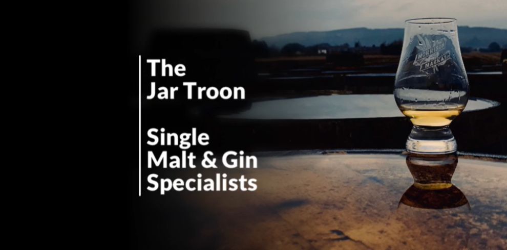 An image of a glass of whiskey with a lovely scenic backdrop. There is text that says 'The Jar Troon, Single Mail & Gin Specialists'.