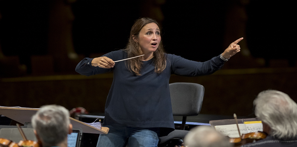 A picture of a conductor leading an orchestra. She stands on a podium with her face to the camera, baton in hand, mid-motion as she directs the musicians.