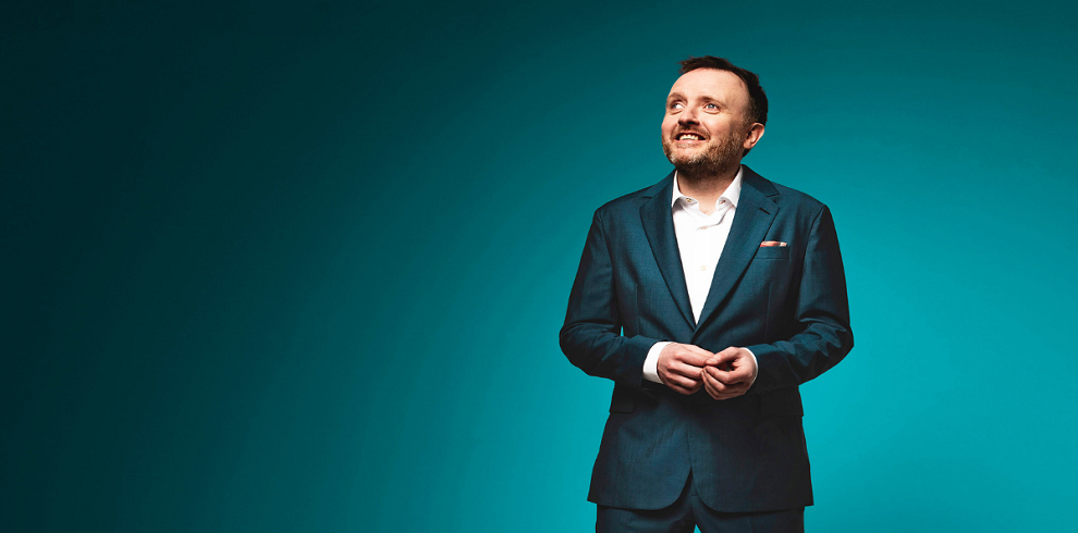 A picture of Chris McCausland standing smiling, dressed in a smart suit.