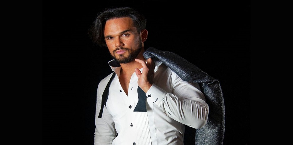 A picture of Gareth Gates, dressed smartly and looking into the camera,, he's holding a jacket over his shoulder and his shirt is open at the top with a bow tie undone.