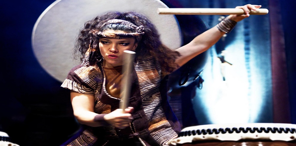 A performer from Mugenkyo Taiko Drummers energetically playing a large taiko drum, wearing traditional costume and headpiece, with dramatic stage lighting in the background.