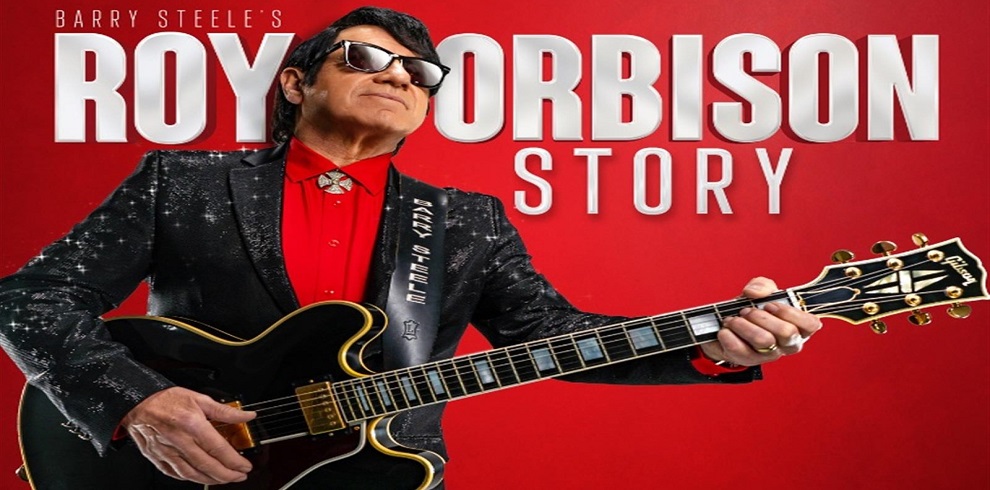 A cast member dressed as Roy Orbison performs on stage, holding a guitar mid-strum. He wears Orbison’s iconic dark sunglasses and a dark suit, reminiscent of the singer’s classic look, with a focused expression that adds to the performance's intensity. The background is softly lit, highlighting the musician while maintaining a retro concert ambiance.