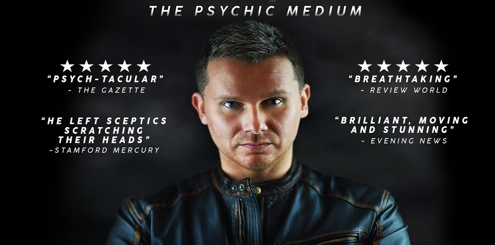 This is an image of a psychic medium, presented with a dramatic, intense look against a dark, misty background. The person is dressed in a dark leather jacket, looking directly into the camera with a serious expression. Above him, the title "The Psychic Medium" is displayed in white, uppercase text. Surrounding him are quotes from various publications with five-star ratings, praising his performances: "Psych-tacular" - The Gazette "He left sceptics scratching their heads" - Stamford Mercury "Breathtaking" - Review World "Brilliant, moving, and stunning" - Evening News The overall design is striking, emphasizing the mystery and allure associated with his psychic abilities.