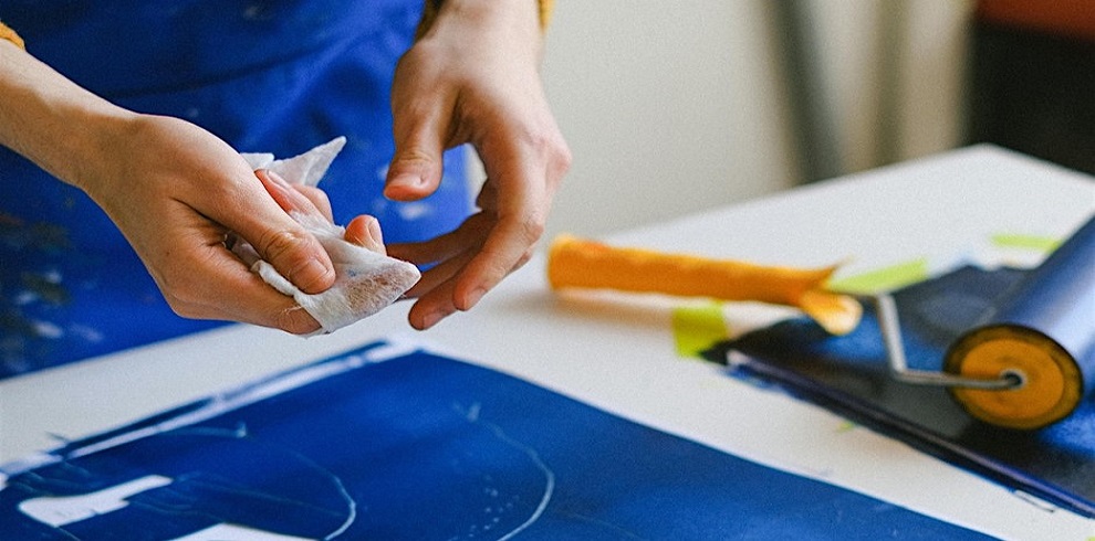 Close-up of hands engaged in a printmaking process, cleaning a tool or surface with a cloth. A brayer (ink roller) and a blue inked plate are visible on a work table, suggesting an art or craft activity.