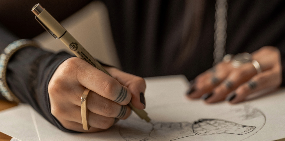 Close-up of a person's hand sketching on paper with a fine-tipped pen, surrounded by a calm and creative atmosphere. The person is wearing stylish rings and has visible tattoos on their fingers, emphasizing individuality and artistic expression. Ideal for promoting a mindfulness workshop focused on creativity and drawing.