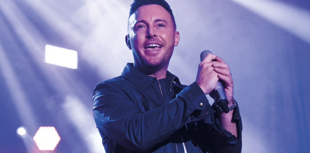 The image shows Nathan Carter performing live at a concert. He is holding a microphone in his hands, smiling warmly, and engaging with the audience. He is dressed in a dark jacket and is illuminated by bright stage lights, creating a vibrant atmosphere. The background features soft, colourful lighting effects, adding to the energy of the performance.