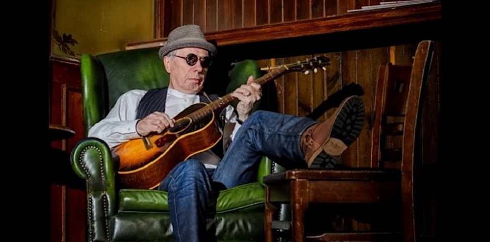 A picture of Al Fleming sitting comfotable on a chair playing his guitar. He is wearing a hat and sun glasses.