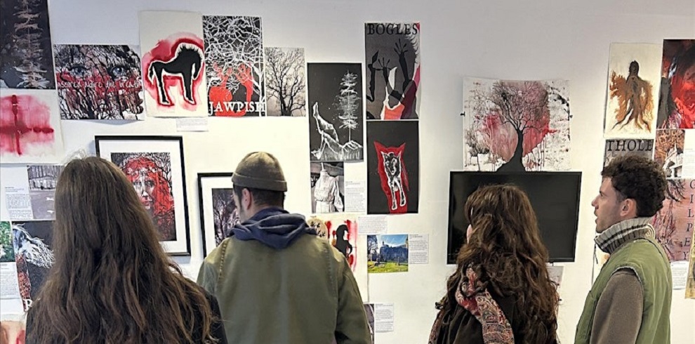 A photograph of four people in an art gallery shows them observing a collection of mixed-media artworks displayed on a white wall. The pieces include illustrations, paintings, and digital prints featuring bold colors, surreal imagery, and text elements, with themes of folklore, mythology, and nature. Some artworks incorporate red and black ink splashes, depictions of animals, trees, and human figures, and words like "Bogles" and "Nawpish." The visitors are dressed in warm clothing, with two wearing beanies and one wrapped in a patterned scarf. A television screen is mounted among the artworks, and printed descriptions are attached to the wall, providing context for the exhibition.