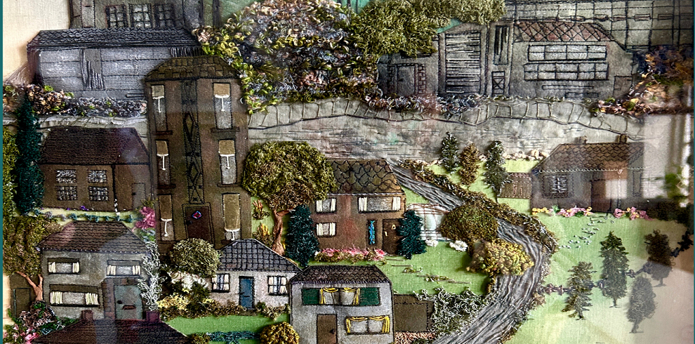 This is a textile and mixed-media artwork depicting a layered townscape with embroidered and painted elements. The artwork features a combination of fabric, thread, and paint to create depth and texture, showcasing houses, trees, and industrial buildings. The foreground includes detailed stitched houses with windows and doors, surrounded by embroidered greenery, bushes, and flowers. A winding road moves through the scene, leading to more buildings in the middle and background, which are drawn and stitched in muted tones. Trees and foliage are embroidered in vibrant greens, purples, and pinks, adding contrast and a three-dimensional effect. The artwork combines realism with an impressionistic texture, highlighting the details of architecture and nature through fabric and threadwork.