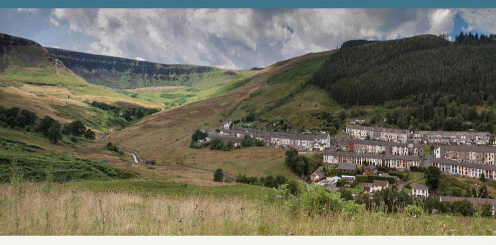 A panoramic view of a former mining village nestled in a lush valley. The village consists of rows of terraced houses stretching along the hillside, with green fields and patches of woodland surrounding the settlement. Rolling hills and a small stream meander through the valley, reflecting the area's natural beauty. The landscape hints at the village's industrial past, now blending into the tranquil countryside.