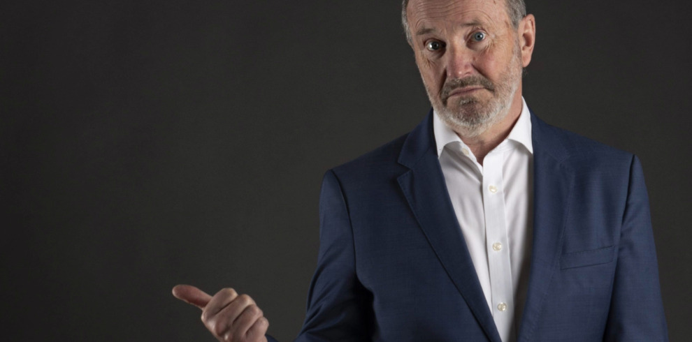 Comedian Fred MacAulay standing against a dark background indoors.