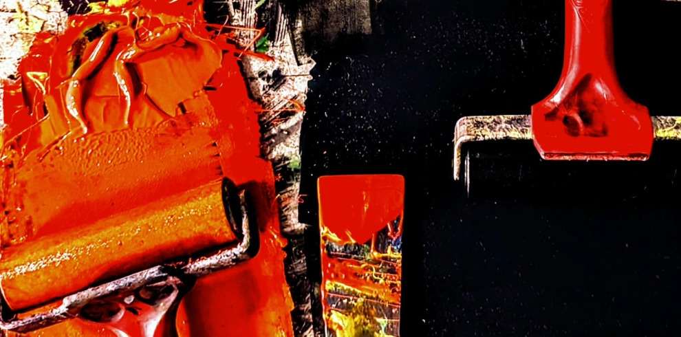 A close-up view of printmaking tools and materials from The Printroom Exhibition. The image features ink rollers covered in thick, vibrant orange ink, contrasting against a black background. One roller is coated with a textured layer of ink, while another is clean with a red handle. A small print with a reflective surface showcases abstract patterns. The composition highlights the bold colors, rich textures, and the hands-on process of printmaking.