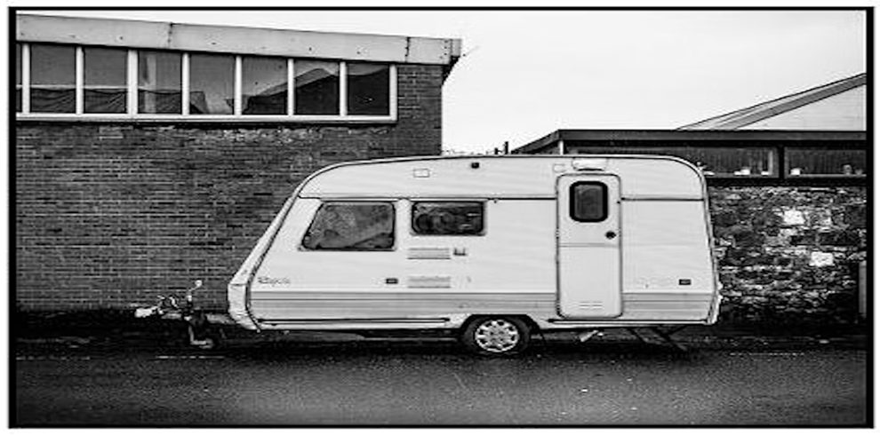 Black and white photograph of a small caravan parked on a street beside a brick industrial building with large windows. A stone wall and another building with a sloped roof are visible in the background. The scene is quiet and slightly overcast, capturing a raw, urban atmosphere.
