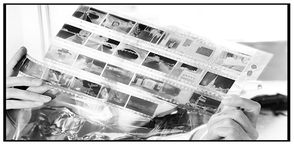 A person holds up a sheet of photographic film negatives encased in a clear protective sleeve. The negatives display various scenes and objects. The image is in black and white, with the person's hands visible grasping the edges of the sleeve, and a soft, bright background suggesting a well-lit room.