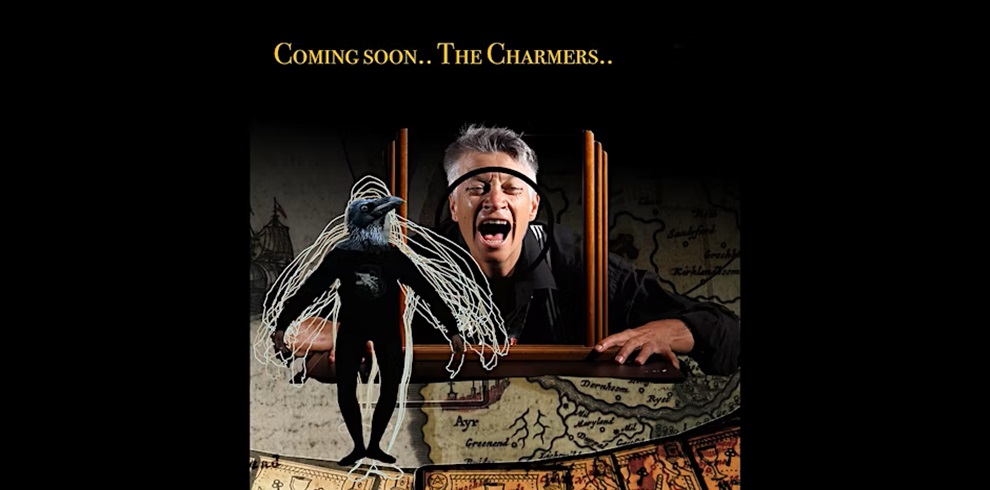 A surreal promotional image featuring a dramatic scene with the text "Coming soon.. The Charmers.." at the top. In the center, a man with gray hair is screaming or acting emotionally while emerging from a wooden picture frame. His head is encircled by a black band, and he leans forward with an intense expression. In front of him is a ghostly, humanoid figure with bird-like features and extended transparent wings, as if it's part of or escaping from the frame. The background includes a vintage map, adding a mysterious, historical atmosphere. The overall tone suggests an upcoming theatrical or artistic performance.