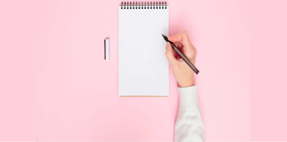A person’s right hand is holding a black pen above a blank white spiral notebook on a pink background. An eraser with a black stripe lies to the left of the notebook. The person is wearing a light-colored long-sleeve shirt.