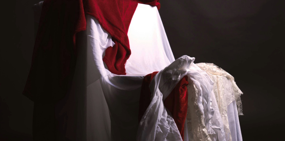 A dramatic still-life photograph of a chair draped in various fabrics. A white cloth covers the chair, with a deep red blanket cascading over the top and side. Additional white and lace textiles are layered over the seat and spill onto the floor. The background is dark, emphasizing the textures and contrast of the fabrics in soft lighting.