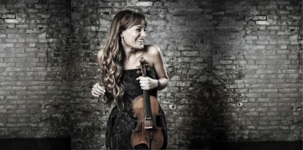 An image of a woman in a black evening gown standing in front of a brick wall holding a violin.
