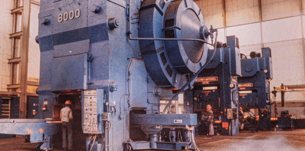 Alt text: Industrial scene inside a large manufacturing facility featuring several massive blue metal stamping presses, each labeled "8000" and "WEILERS & MITCHELL." Workers in protective gear stand at the machines, highlighting the scale of the equipment. The facility has high ceilings and ample lighting, emphasizing its spacious and industrial environment.