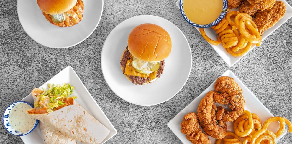An overhead shot shows four white square plates with various food items on a speckled gray surface. In the center, a plate holds a classic smash burger with melted cheese and pickles on a bun. To its upper left, another plate features a similar burger, partially visible. On the lower left, a plate presents two wraps with lettuce and sauce, accompanied by a small blue bowl of white dip. The plate on the upper right contains a generous portion of fried chicken strips and curly fries, along with a small blue bowl of golden cheese sauce. The plate on the lower right also has fried chicken and curly fries.