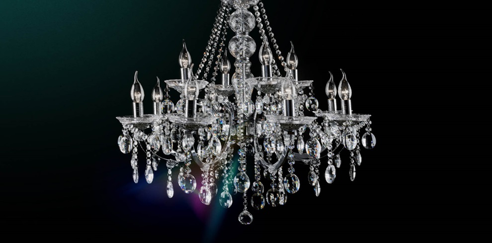 An ornate crystal chandelier with multiple arms, each holding a candle-shaped light bulb. The chandelier is decorated with hanging crystal prisms and beads that reflect light, creating a sparkling effect. The dark background enhances the chandelier's brilliance.
