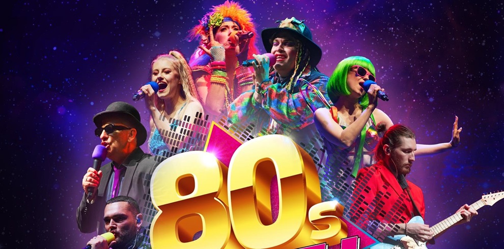 A vibrant stage scene featuring six performers in 1980s-themed costumes, standing in front of a starry background. Large, bold yellow text reading '80s' is prominently displayed in front of them. The performers, whose faces are pixelated for privacy, are holding microphones and musical instruments, including an electric guitar.