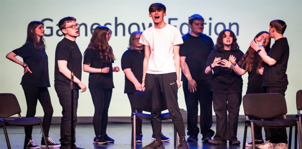 A group of young performers are on stage in a theatrical scene, wearing casual black outfits, except for one person in the center who stands out in a white t-shirt. The backdrop displays the words “Gameshow Edition.” The performers express various emotions through their body language and facial expressions, suggesting a dramatic or comedic performance. Several chairs are scattered across the stage, adding to the theatrical setup.