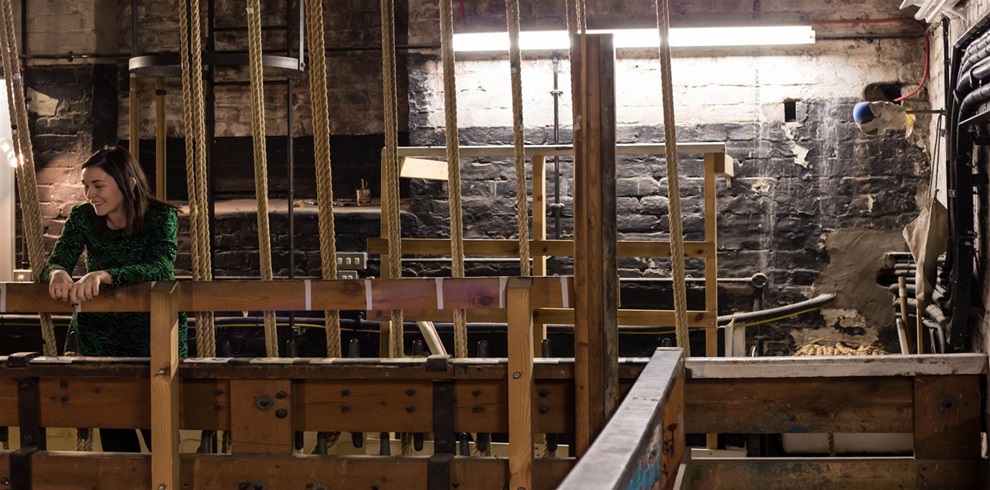 A woman in a green patterned dress stands smiling behind a wooden railing in a backstage area of a theater. Around her are numerous vertical ropes used for stage rigging, along with wooden and metal structures. The brick wall in the background is painted black and appears aged, with patches of peeling paint and exposed plaster. A fluorescent light fixture above casts a soft glow over the scene, highlighting the industrial and historic feel of the space
