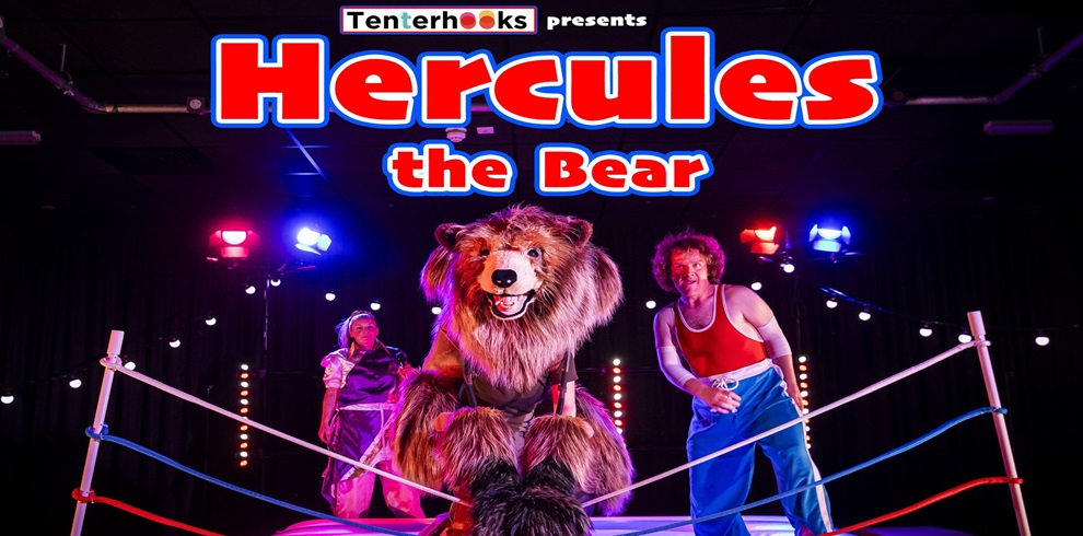 A theatrical promotional image for "Hercules the Bear," presented by Tenterhooks. The scene features a person in a large, realistic bear costume standing in a boxing ring, flanked by two actors dressed in colorful 1980s-style athletic outfits. Bright stage lights illuminate the background, creating a vibrant and dramatic atmosphere. The title "Hercules the Bear" is displayed in bold red and blue letters above the characters.