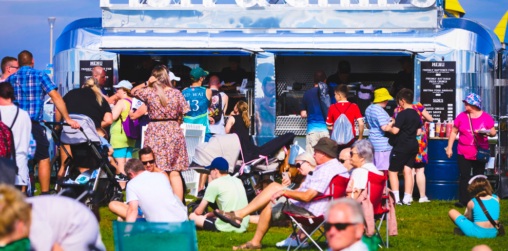 A crowd of people standing and sitting on the grass outside a mobile food truck.
