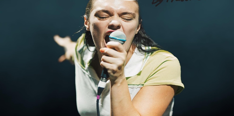 A person is performing on stage, holding a microphone close to their mouth with one hand while extending the other arm outward. The person's face is blurred. They are wearing a light-colored shirt with green accents. The background is dark and out of focus, emphasizing the performer.