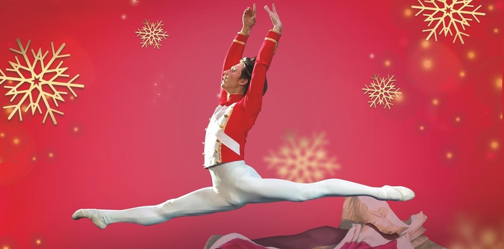 A ballet dancer mid-leap against a festive red background with golden snowflakes. The dancer wears a red and white Nutcracker-style costume, white tights, and ballet shoes. Arms are raised overhead and legs extended in a split. The dancer's face is obscured by a square.