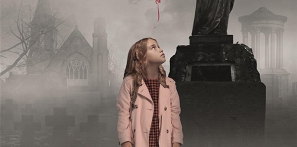 The image shows a young child standing in front of a statue in what appears to be a graveyard. The child's face is obscured by a square, and the child is wearing a pink coat over a red and black checkered dress. The background includes gravestones, an old church with gothic architecture, and another building with columns partially visible through the fog. A red balloon is floating above the child's head, tied to the child's wrist.