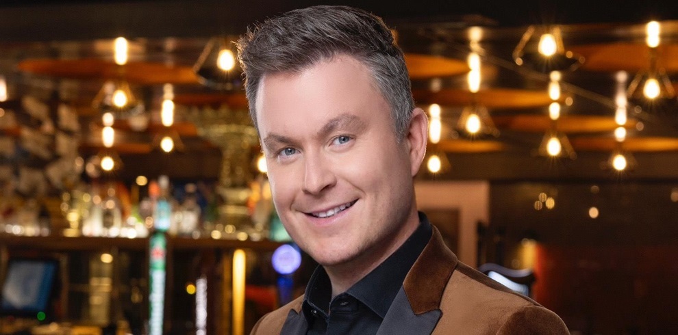 The image shows a person with a blurred face standing in what appears to be a bar or restaurant setting. The background includes hanging lights, bottles on shelves, and a bar counter. The person is wearing a dark shirt and a brown jacket.