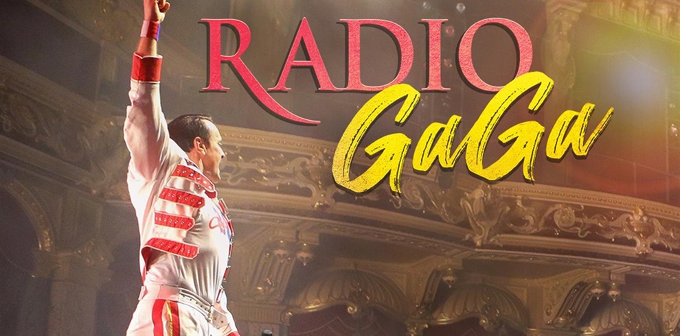 A performer dressed in a white and red costume reminiscent of Freddie Mercury raises a triumphant fist on stage in an ornate theatre setting. The bold title "RADIO GaGa" appears beside him, with "RADIO" in red uppercase letters and "GaGa" in a dynamic yellow handwritten style. The scene evokes the energy and spectacle of a Queen tribute concert.